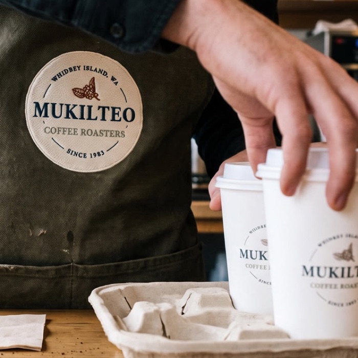 The Mukilteo roundel embroidered on a canvas apron, with two branded cups and a carry tray