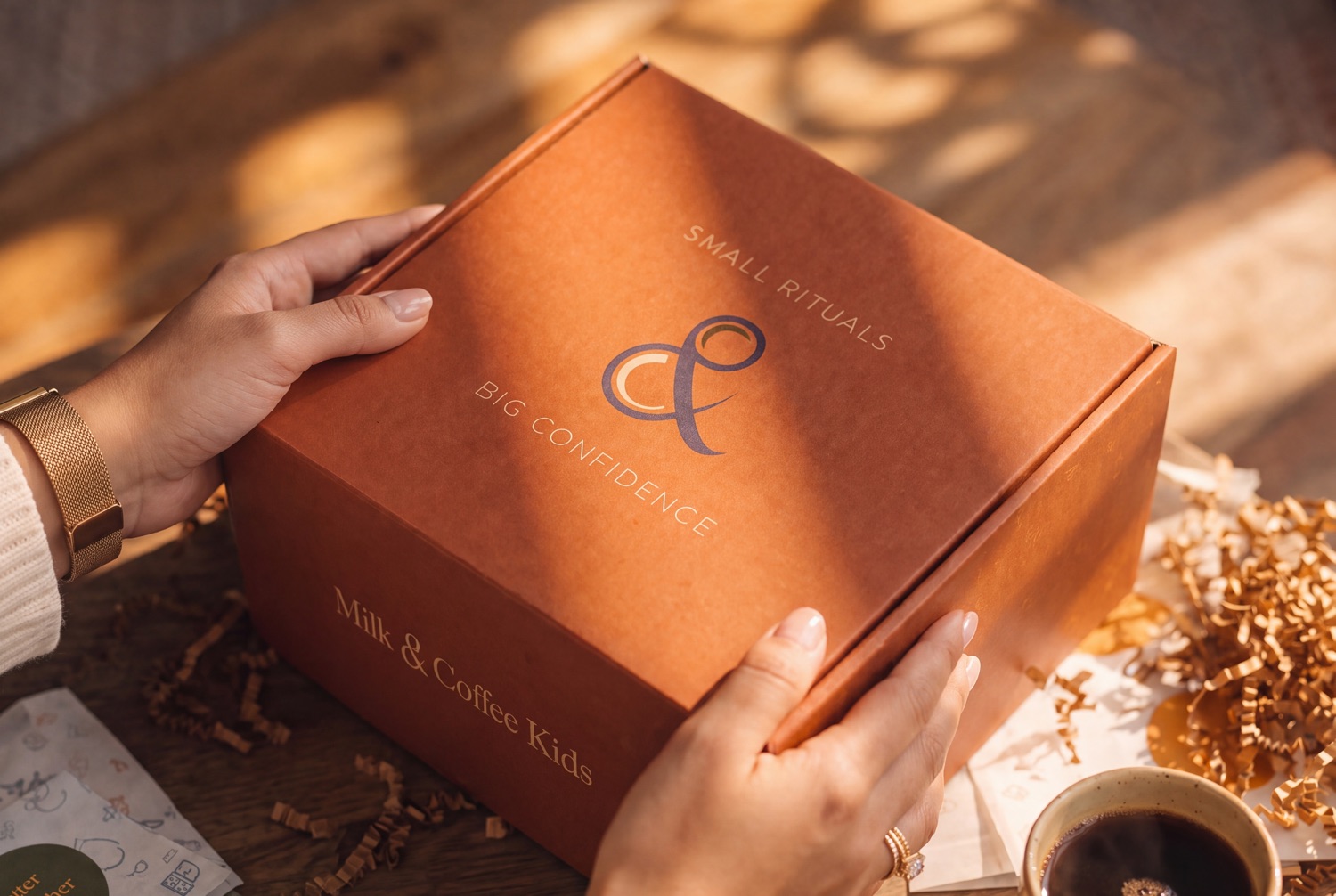 The closed mailer held over the table, the ampersand mark and both lines of the lockup printed across the lid, cards and a coffee beside it