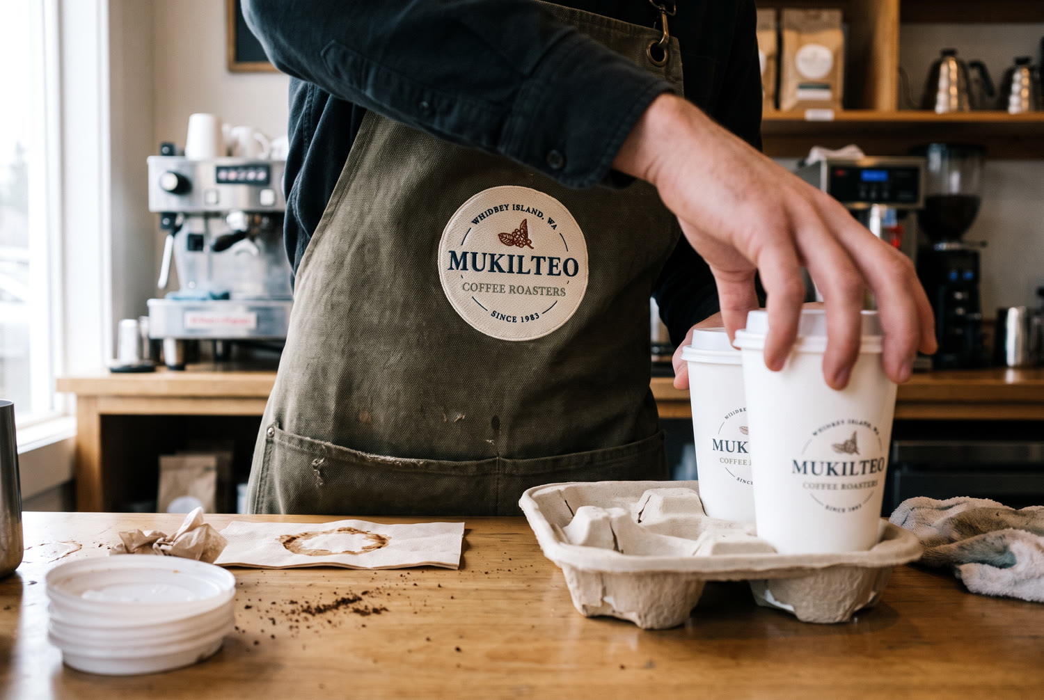 A barista in a canvas apron carrying the embroidered Mukilteo badge, setting two branded cups into a carry tray